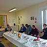 bottles, conversation, cups, dining_room, dog, exit_sign, food, group, indoor, juice, kitchen, people, plates, sandwiches, seated, smartphone, snacks, standing, table, window