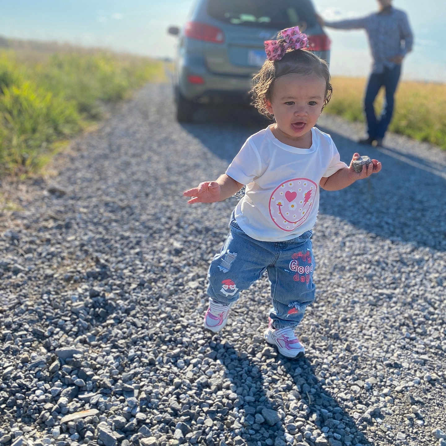 Khilynn joined the competition — help win amazing prizes! background, blue_sky, car, casual, child, grass, gravel_road, jeans, man, nature, outdoor, person, pink_bow, rock, shadow, sneakers, sunny_day, toddler, walking, white_tshirt