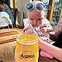 baby, sunglasses, glass, cider, table, tattoo, arm, indoor, window, person, wood, drink, curious, child, pink_clothing, highchair, lamp, cozy, cafe, reflection