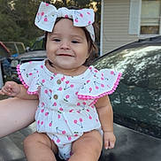 Eliana is registered to the contest to win money with this photo: baby, child, infant, smiling, portrait, outdoor, car, vehicle, bow, hair_bow, dress, floral_pattern, cherry_pattern, pink, seated, barefoot, hand, house, trees, daylight