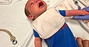 Adrian is registered to the contest to win money with this photo: baby, bed, blue_onesie, cabinet, care, clinic, crying, gloved_hand, healthcare_worker, hospital, hospital_room, infant, medical, medical_procedure, medical_supplies, newborn, patient, person, syringe, white_bed_sheet