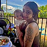 baby, woman, tattoo, outdoor, smiling, holding, food, plate, car_seat, toy, table, sky, cloud, greenery, railing, person, portrait, casual, daylight, happy