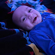 Toa-Malosi is registered to the contest to win money with this photo: baby, blanket, blue_clothing, child, closeup, cozy, cute, dimples, face, happy, indoor, infant, lying_down, patterned_blanket, person, portrait, sleepwear, smiling, soft_lighting, warmth