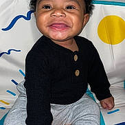 Amarjae is registered to the contest to win money with this photo: baby, smiling, child, sitting, black_sweater, gray_pants, curly_hair, play_mat, colorful_background, happy, indoors, cute, portrait, young_child, person, infant, one_person, bright, cozy, buttoned_clothing