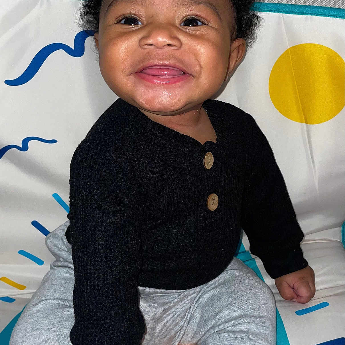 Amarjae is registered to the contest to win money with this photo: baby, smiling, child, sitting, black_sweater, gray_pants, curly_hair, play_mat, colorful_background, happy, indoors, cute, portrait, young_child, person, infant, one_person, bright, cozy, buttoned_clothing