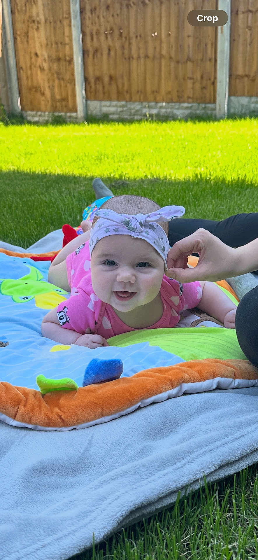 Ayla is registered to the contest to win money with this photo: baby, smiling, headband, pink_outfit, blanket, grass, outdoor, hand, fence, sunlight, infant, cute, happy, tummy_time, colorful, toy, head, face, person, nature