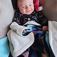 Liam is registered to the contest to win money with this photo: baby, blanket, black_hoodie, pink_fluffy_accessory, smiling, face, child, cozy, soft_texture, indoor, couch, comfortable, cute, portrait, infant, warm, happy, clothing, relaxed, wrapped