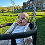 Isaac-George is registered to the contest to win money with this photo: baby, child, swing, playground, outdoor, smiling, happy, grass, park, daytime, blue_sky, chain, fence, house, residential, casual_clothing, vest, person, infant, recreation