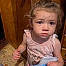 child, toddler, indoor, curly_hair, snack, bag, wooden_door, cabinet, flooring, patterned_floor, pink_shirt, curious, blurry, person, young_child, standing, holding, expression, home, candid
