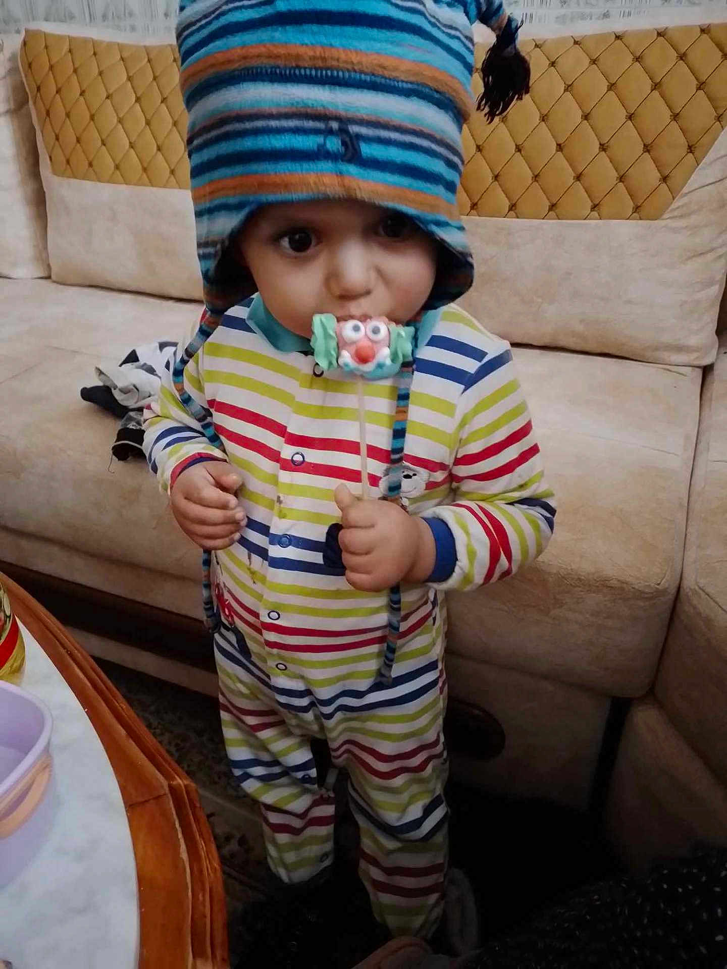 Ilyes participe au concours pour gagner de l'argent avec cette photo : toddler, child, striped_clothing, hat, indoor, lollipop, candy, couch, furniture, person, face, eyes, hand, table, colorful, striped, cute, winter_hat, playful, standing