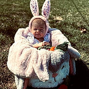 Brooklynn is registered to the contest to win money with this photo: baby, bunny_ears, plush_blanket, carrot_toy, outdoor, grass, sunlight, cute, child, infant, costume, headwear, toy, soft_texture, nature, portrait, sitting, expression, warm_clothing, animal_theme