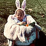 baby, bunny_ears, plush_blanket, carrot_toy, outdoor, grass, sunlight, cute, child, infant, costume, headwear, toy, soft_texture, nature, portrait, sitting, expression, warm_clothing, animal_theme