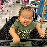 Nelson is registered to the contest to win money with this photo: toddler, child, shopping_cart, store, aisle, smile, curly_hair, green_clothing, baby, person, indoor, retail, grip, happy, cute, young_child, playful, shopping, black_cart, mischievous