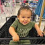 toddler, child, shopping_cart, store, aisle, smile, curly_hair, green_clothing, baby, person, indoor, retail, grip, happy, cute, young_child, playful, shopping, black_cart, mischievous