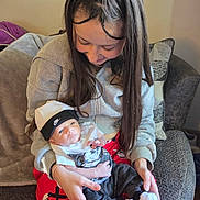 Lucas is registered to the contest to win money with this photo: baby, casual, child, clothing, couch, family, girl, gray_hoodie, hat, holding, home, indoor, infant, long_hair, love, person, pillow, red_pants, seated, socks