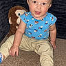 baby, beige_pants, blue_shirt, carpet, casual_clothing, child, cute, happy, home, indoor, monkey, person, playful, plush_toy, portrait, seated, smiling, socks, toddler, young_child