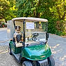 dog, australian_shepherd, golf_cart, vehicle, outdoor, parked, trees, foliage, road, driveway, sunlight, green, long_hair, floppy_ears, tongue_out, sitting, pet, transportation, windshield, funny