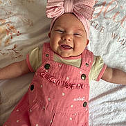 Kylie is registered to the contest to win money with this photo: baby, smiling, pink_bow, headband, pink_overalls, infant, happy, lying_down, blanket, floral_pattern, cute, child, portrait, soft_lighting, indoors, clothing, baby_girl, face, toddler, apparel