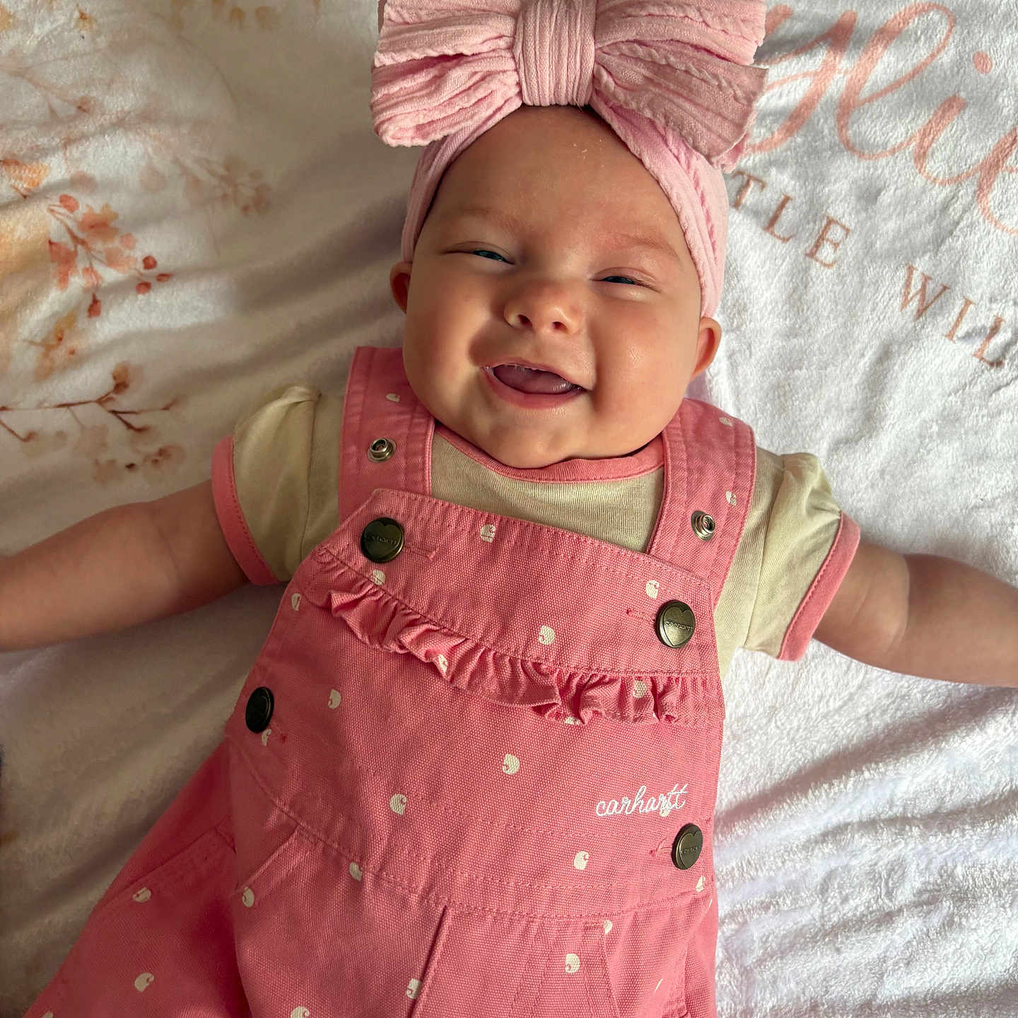 Kylie is registered to the contest to win money with this photo: baby, smiling, pink_bow, headband, pink_overalls, infant, happy, lying_down, blanket, floral_pattern, cute, child, portrait, soft_lighting, indoors, clothing, baby_girl, face, toddler, apparel