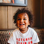 Noah joined the competition — help win amazing prizes! toddler, child, smiling, curly_hair, white_shirt, text_on_shirt, ketchup, stain, indoor, bench, patterned_bench, happy, portrait, face, cute, young_child, casual_clothing, warm_lighting, person, seated