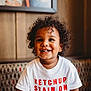 toddler, child, smiling, curly_hair, white_shirt, text_on_shirt, ketchup, stain, indoor, bench, patterned_bench, happy, portrait, face, cute, young_child, casual_clothing, warm_lighting, person, seated
