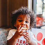 Noah is registered to the contest to win money with this photo: blurred_background, booth, child, curly_hair, cute, diner, eating, face, food, hands, happy, indoor, person, portrait, red_straw, snack, table, toddler, white_shirt, window