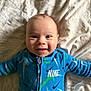 baby, smiling, blue_clothing, nike, onesie, infant, lying_down, blanket, happy, cute, child, face, portrait, indoors, soft_texture, patterned, young, person, casual, closeup