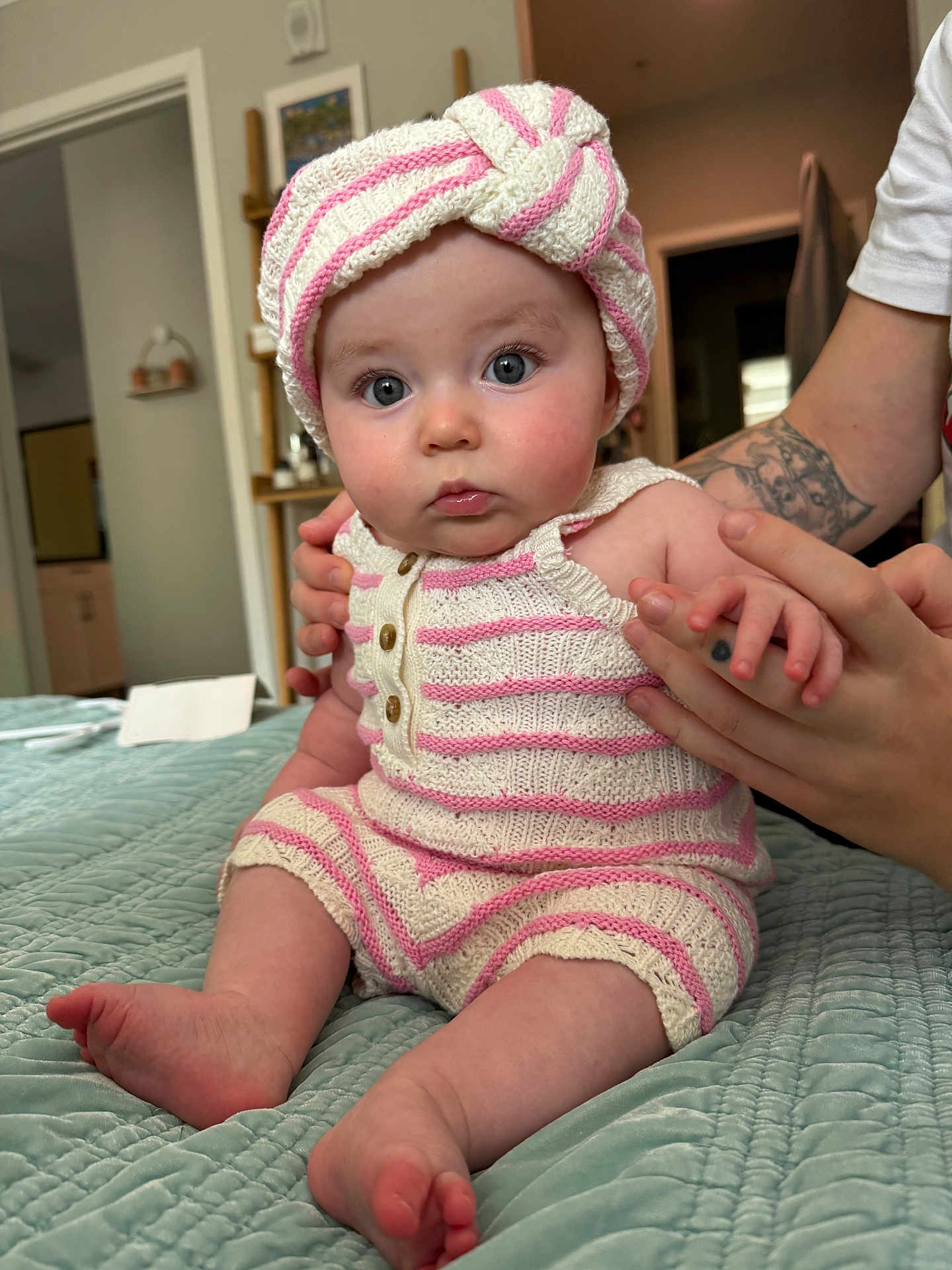 Salem is registered to the contest to win money with this photo: baby, infant, knitwear, striped_clothing, turban, hand, tattoo, bed, quilt, indoor, person, cute, child, blue_eyes, soft_lighting, portrait, skin, sitting, supporting_hand, cozy