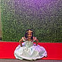 celebration, child, cute, event, fashion, floor, formal_wear, girl, green_hedge, hair_beads, happy, indoor, person, portrait, princess_dress, red_carpet, sitting, smiling, tiara, young