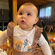 Bertille a rejoint le concours — aidez-le/la à gagner de superbes lots ! baby, infant, child, face, big_eyes, cheeks, curious_gaze, ruffled_clothing, suspenders, onesie, indoor, living_room, wooden_beam, curtains, natural_light, play_gym, toy, hand, seated, portrait