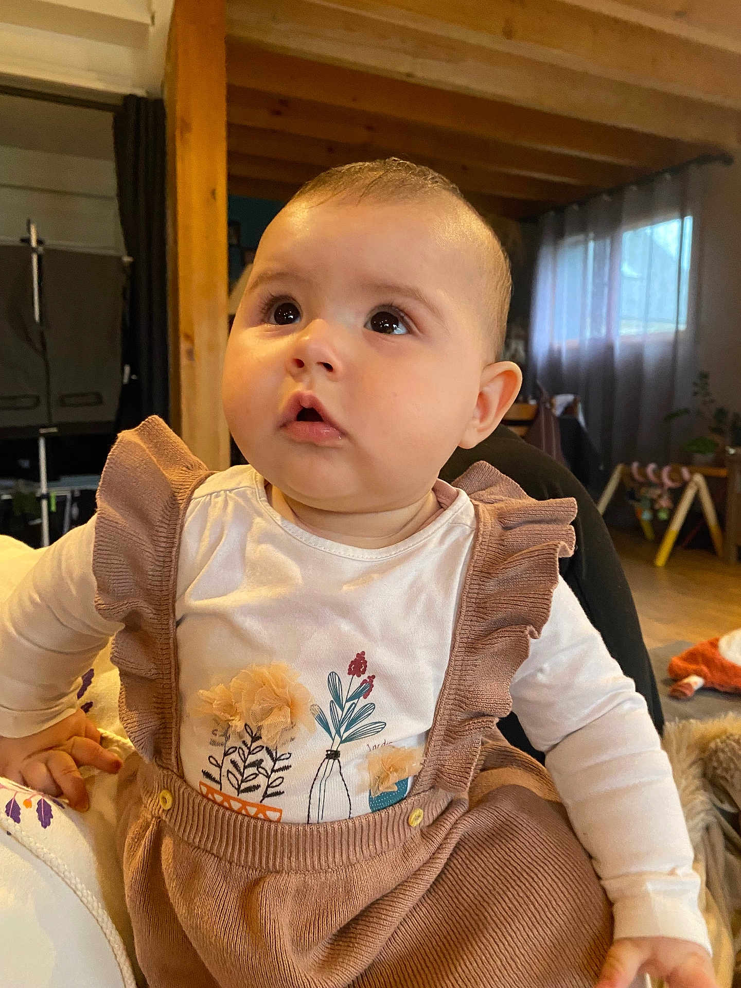 Bertille a rejoint le concours — aidez-le/la à gagner de superbes lots ! baby, infant, child, face, big_eyes, cheeks, curious_gaze, ruffled_clothing, suspenders, onesie, indoor, living_room, wooden_beam, curtains, natural_light, play_gym, toy, hand, seated, portrait