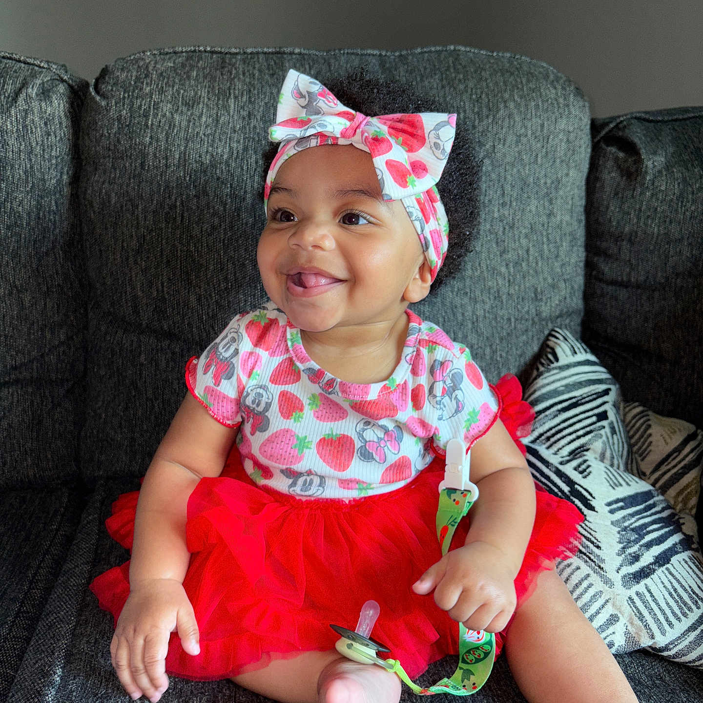 Malaya is registered to the contest to win money with this photo: baby, bow, bright_colors, child, couch, cute, funny_face, happy, headband, indoor, infant, pacifier, patterned_clothing, pillow, portrait, red_skirt, sitting, smile, toddler, tongue_out