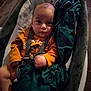baby, baby_seat, blanket, child, comfort, cozy, expression, face, floor, green_blanket, indoor, infant, looking_at_camera, orange_shirt, person, portrait, seated, straps, wooden_floor, young_child
