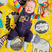Dougie is registered to the contest to win money with this photo: baby, infant, smiling, colorful_clothing, playmat, yellow_background, striped_shirt, overalls, blue_socks, child, happy, toy, mirror, elephant, lion, octopus, zebra, raised_arms, indoor, cute