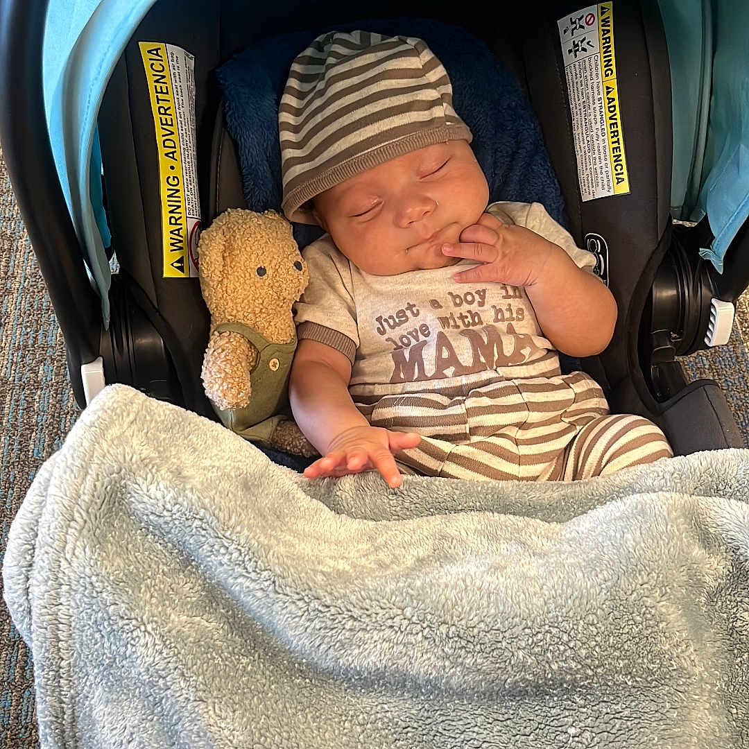 Luis joined the competition — help win amazing prizes! baby, sleeping, car_seat, plush_toy, blanket, striped_clothing, hat, footwear, floor, person, infant, cozy, child, cute, indoors, soft_texture, resting, napping, toddler, peaceful