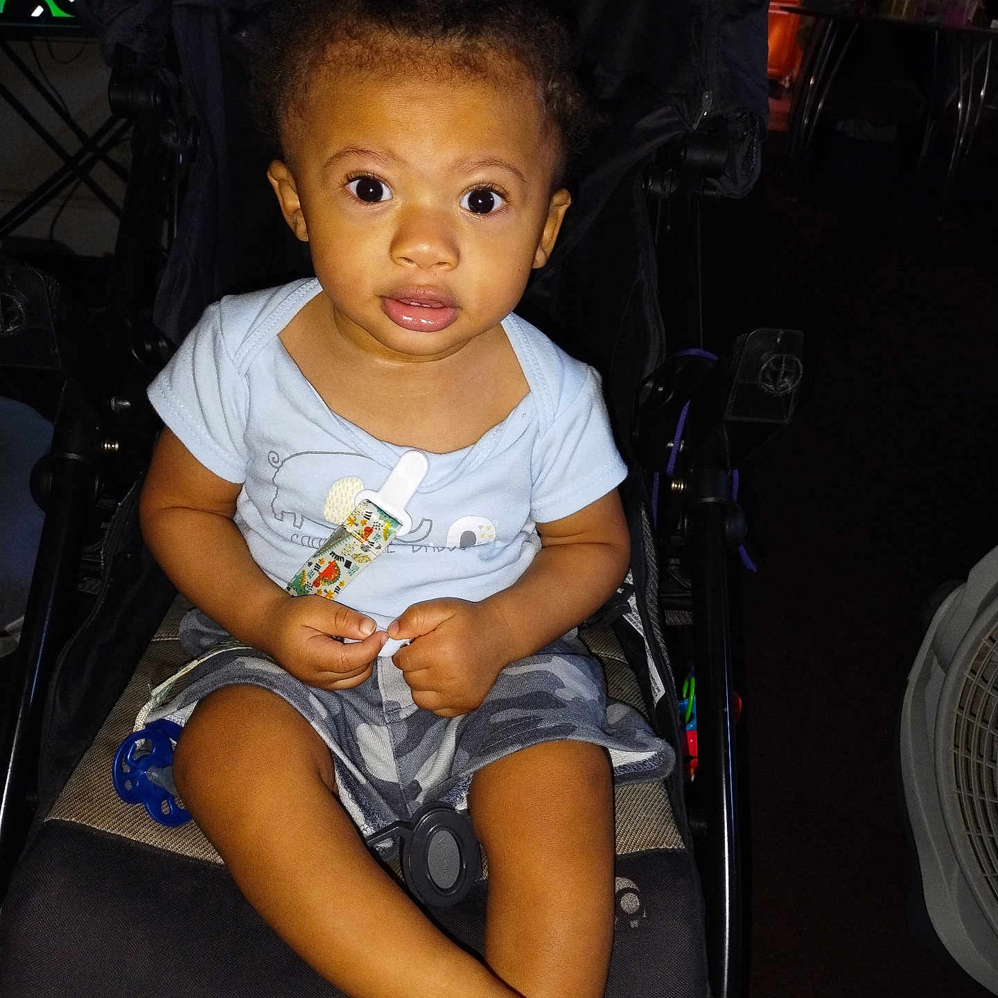 Elijah is registered to the contest to win money with this photo: baby, barefoot, blue_shirt, casual_clothing, child, comfort, curly_hair, expression, face, indoor, infant, light, pacifier, person, seat, shorts, stroller, toddler, toy, young_child