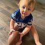 toddler, child, football, blue_jersey, indoor, wooden_floor, barefoot, curious, blonde_hair, playing, sport, young_child, casual, sitting, looking_up, hands, feet, toy, closeup, portrait