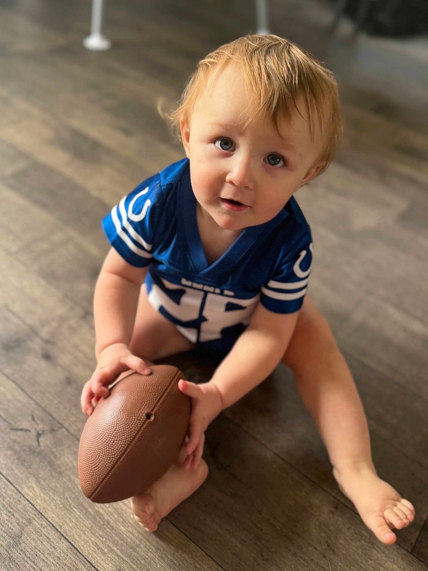 Asher is registered to the contest to win money with this photo: toddler, child, football, blue_jersey, indoor, wooden_floor, barefoot, curious, blonde_hair, playing, sport, young_child, casual, sitting, looking_up, hands, feet, toy, closeup, portrait