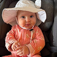 Magdalene is registered to the contest to win money with this photo: baby, infant, child, car_seat, hat, pink_clothing, onesie, floral_pattern, cute, smiling, indoors, portrait, seatbelt, headwear, small_hands, young_child, face, eyes, expression, cozy
