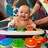Magdalene is registered to the contest to win money with this photo: baby, smile, toy_piano, colorful, playtime, infant, hanging_toys, adult_support, indoor, bedroom, happy, child, clothing, person, hands, face, head, furniture, blanket, cute