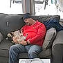 beanie, casual_clothing, couch, cozy, dog, gray_couch, home, indoor, living_room, man, napping, pet, pillows, red_shirt, relaxing, resting, seated, sleeping, window, winter
