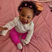 Island is registered to the contest to win money with this photo: baby, child, smiling, curly_hair, pink_clothing, white_top, sitting, bed, blanket, hand_holding, happy, infant, cute, indoor, person, portrait, pink_bedspread, hair_ties, young_child, cozy