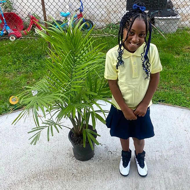 Skylar is registered to the contest to win money with this photo: bicycle, braid, child, clothing, cornrows, face, female, girl, hair, head, outdoors, person, plant, sitting, skirt, sleeve, standing, student, transportation, vehicle