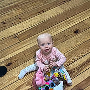 Paisley joined the competition — help win amazing prizes! baby, child, pink_clothing, wooden_floor, toys, bowl, plush_toy, sitting, indoor, playtime, cute, smiling, colorful, person, sock, flooring, object, happy, looking_up, infant