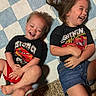 Titus is registered to the contest to win money with this photo: child, laughing, toddler, young_child, smiling, black_tshirt, red_shorts, denim_shorts, car_print, lying_down, blue_mat, patterned_mat, indoor, happy, sibling, playful, barefoot, hands, hair, joy