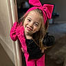 child, girl, smiling, pink_bow, pink_jacket, pink_pants, black_top, long_hair, indoor, door_frame, playful, happy, portrait, young_child, casual_clothing, person, fashion, cute, standing, light