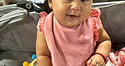 Mckenzie is registered to the contest to win money with this photo: baby, backyard, bib, bracelet, brick_wall, brown_hair, child, closeup, dress, earring, facial_expression, infant, outdoor, pink_clothing, portrait, sitting, smiling, stroller, toy, wooden_fence