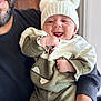 Ayaan is registered to the contest to win money with this photo: baby, infant, smiling, happy, hat, knit_hat, pom_poms, green_clothing, white_clothing, adult, arm, holding, portrait, indoor, cozy, cute, child, person, face, close_up