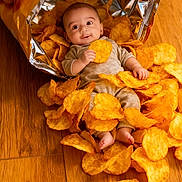 Ayaan is registered to the contest to win money with this photo: baby, infant, potato_chips, lays, snack, food, bag, floor, wooden_floor, smiling, cute, child, small, holding, chips, inside, open_bag, happy, playful, indoors