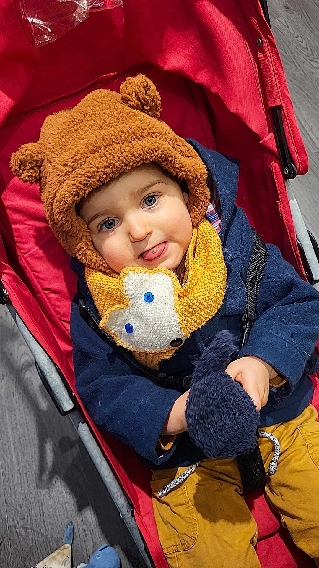 Antonin participe au concours pour gagner de l'argent avec cette photo : baby, bear_hat, blue_eyes, button, child, coat, fabric, hand, hat, plush, portrait, red, scarf, seat, smile, stroller, tongue_out, toy, wood_floor, yellow_pants